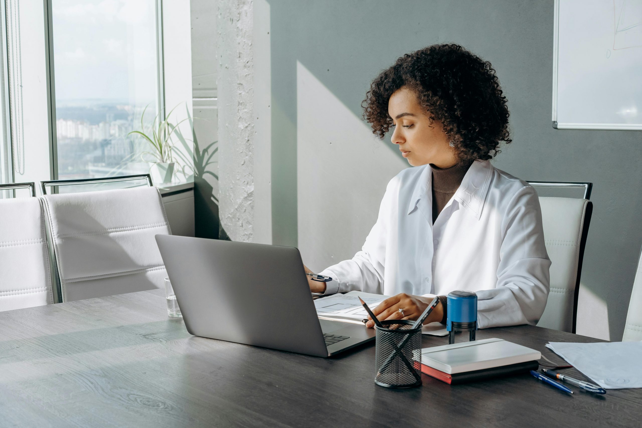 woman at a desk with a laptop, documents, and a notebook in a sunlit room