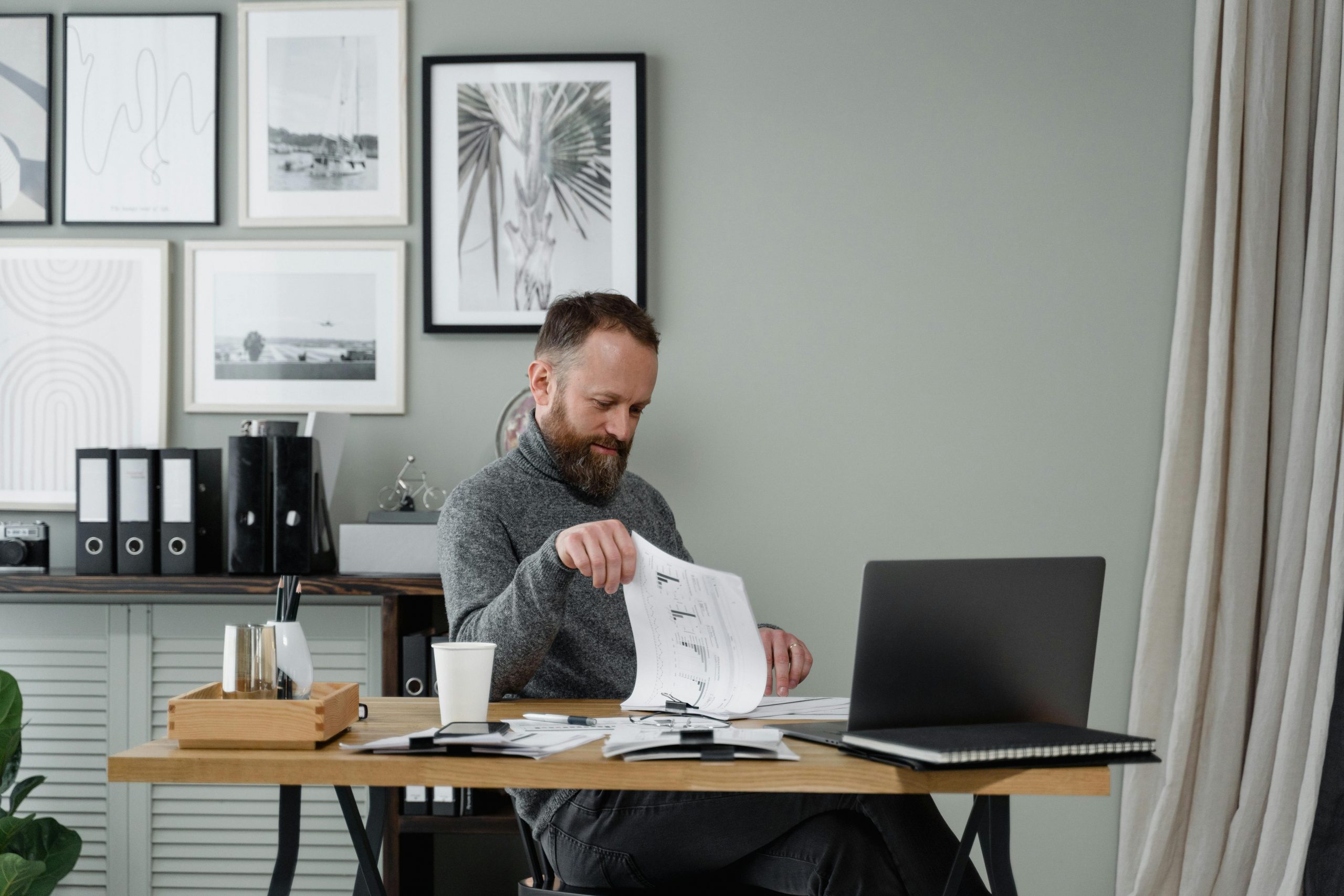 man wearing a gray turtleneck reading a report at a desk with a laptop
